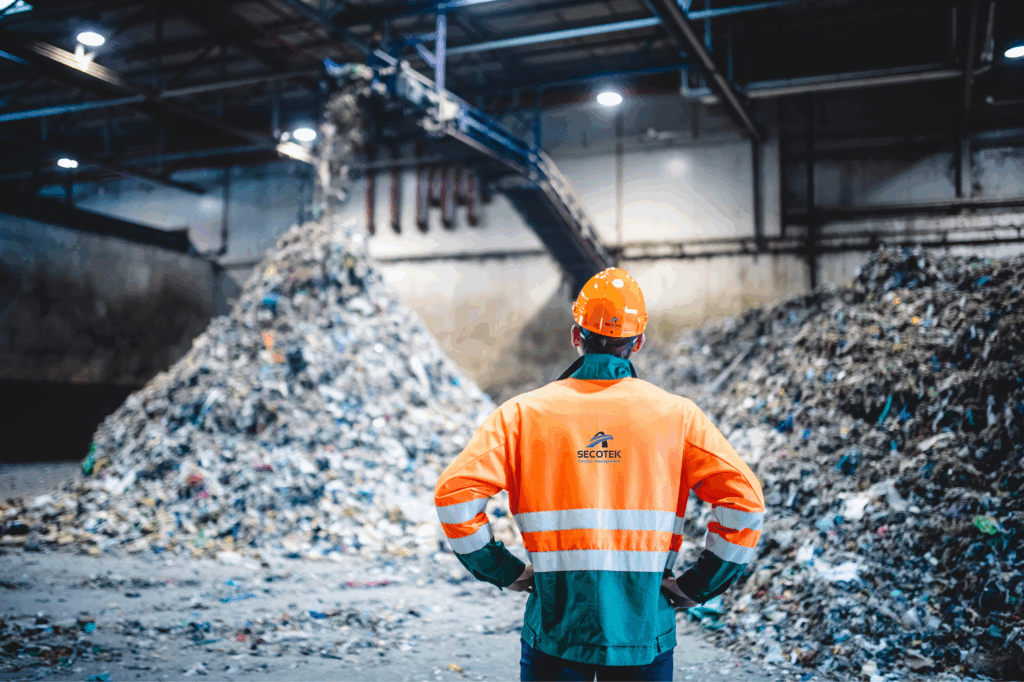 Rückansicht eines jungen männlichen Arbeiters mit Helm, Atemschutzmaske und reflektierender Kleidung, der beobachtet, wie Abfall vom Förderband auf einen Haufen in einer Anlage fällt. unsere service: Entsorgungsdienste Düsseldorf